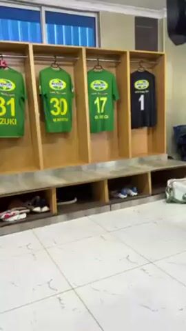 Here is a forensic description of the video:

**Setting:** The video is taken in a locker room. The room is tiled with white and grey square tiles. Along the wall are open-faced wooden lockers. 

**Objects:** Inside each locker hangs a sports jersey. Most of the jerseys are green with a round emblem and a number on the back, along with a name printed below the number. One jersey is black with a number 1 on the back. The numbers on the green jerseys vary, including 9, 11, 17, 18, 21, 27, 30, 31, 33. Below the hanging jerseys, the lockers have shelves containing shoes, sandals, and water bottles. On the floor, several pairs of sports shoes and sandals are scattered.

**Action:** The camera slowly pans along the row of lockers, showcasing the jerseys and the contents of the shelves below.