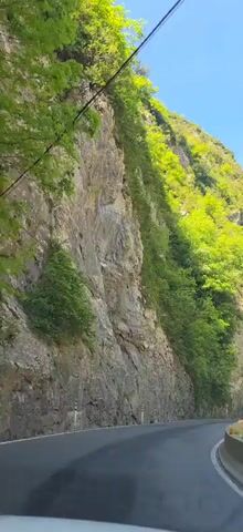 A car travels on an asphalt road through a mountain pass. The road is bordered by a concrete barrier on the right. On either side of the road are steep, rocky cliffs covered in green vegetation. The sky above is clear and blue. The camera is in motion, providing a driver's perspective.