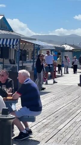 The video shows a sunny day on a wooden pier lined with blue and white striped awnings. People are walking along the pier, and some are sitting at picnic tables. A woman with long dark hair is standing and singing into a microphone. A man sitting at a table to the left of the singer is wearing sunglasses. There are mountains in the distance. A seagull flies by in the background.