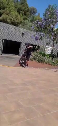 In the first clip, a person is seen skateboarding on a yellow-lined concrete area in front of a building with a textured grey facade. The person is wearing a dark t-shirt, dark shorts, and a baseball cap, and is in motion, appearing to be in the middle of a trick with one leg extended. In the second clip, the same person, now from behind, is walking away from the camera on the same paved area. They are wearing a black tank top, dark shorts, and a tool belt, with green sneakers. A sign with yellow text and a graphic is visible on the building behind them.