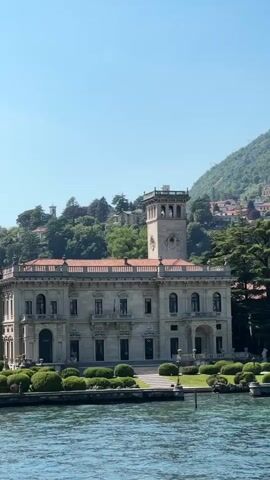 The video captures a scenic view of a large, light-colored building with a distinctive tower, possibly a mansion or villa, situated along the waterfront. The building is adorned with architectural details, including windows, balconies, and decorative elements. The waterfront is lined with meticulously trimmed bushes and a low stone wall. The water is slightly choppy, reflecting the daylight. In the background, a lush green hillside rises, dotted with houses and trees. A smaller structure with white columns and a series of light-colored canopies is visible along the shoreline. The sky is a clear, light blue, suggesting a bright sunny day. The camera appears to be positioned on a boat or other watercraft, offering a dynamic perspective of the landscape.