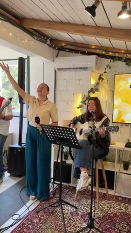 A group of young people are performing music in what appears to be a small, casual venue. There are two women in the center. One woman, standing, sings into a microphone while raising one arm into the air. She is wearing a short-sleeved beige shirt with high-waisted teal pants and is barefoot. The other woman is sitting on a stool and playing an acoustic guitar. She is wearing jeans, white sneakers, and a plaid shirt over a white t-shirt. A third person, a man partially visible on the left, is playing a stringed instrument. There's a music stand with sheet music in front of the women. Behind them, a TV screen displays yellow graphics. The room features rustic decor with exposed wooden beams, string lights, and plants. The floor is covered with a patterned rug. There's a speaker on the floor to the left of the women and an air conditioning unit on the white brick wall in the background.