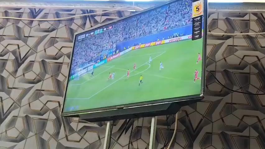 A wide-angle shot shows a television screen displaying a soccer match. The screen is mounted on a wall with a patterned wallpaper. On the television, players in red and white uniforms are on a green soccer field. Spectators are visible in the background. The top right corner of the screen shows a score of 5. The bottom of the television has a black soundbar. The lighting is bright.