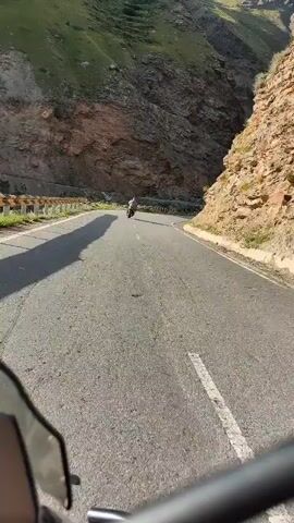 The video shows a first-person perspective of someone riding a motorcycle on a winding, paved mountain road. The road has a white dashed line down the center and a guardrail on the left side in some sections. Steep, rocky, and vegetated mountains rise on both sides of the road. There is another motorcycle rider ahead on the road. The sky appears clear and the lighting suggests it is daytime.