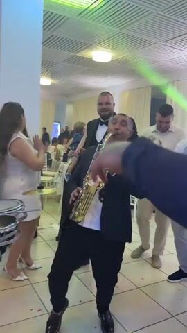 The video shows a wedding reception. A man in a black suit and white shirt is playing a saxophone, and a drummer is to his left. Several guests, mostly women dressed in white or black dresses, are dancing and cheering. A man in a black suit raises his arm in the air. The room is decorated with white tablecloths and white curtains, and there is a disco light.
