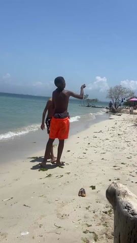 The video captures a bright, sunny day at the beach. Two young, shirtless boys stand on the sandy shoreline. One boy, wearing orange shorts, is holding a kite string, controlling a small, star-shaped kite in the clear blue sky. The other boy, in patterned black and red shorts, stands beside him. Behind them, the beach is dotted with colorful beach umbrellas and lounge chairs. Several people are visible further down the beach. The camera pans up to show the kite in the sky and then back down to the boys on the beach. The sea is calm and turquoise.