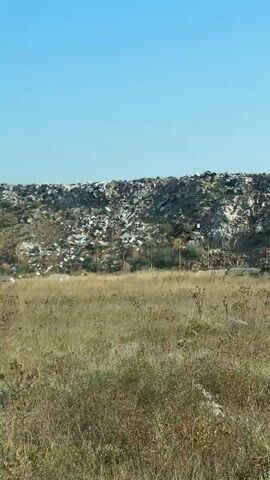 A video shows a large landfill with a clear blue sky in the background. In the foreground, there is dry grass and some vegetation. The landfill is a massive pile of trash, with a lot of white plastic bags and other refuse visible. There are birds flying around the landfill, some of which appear to be seagulls. The area is fenced off with metal posts and some concrete structures. The overall impression is one of a large-scale waste disposal site.