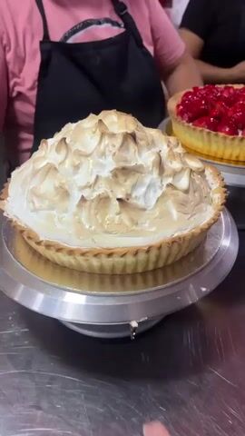 A close-up shot of a meringue-topped tart is in the foreground. Behind it, a strawberry tart is displayed on a silver cake stand. In the background, a group of women wearing aprons and hairnets are visible. One woman on the right is holding a phone and taking a picture of the tarts. Another woman to her left is also holding a phone, possibly taking a picture. A third woman in the background is wearing a pink shirt and an apron. The environment appears to be a bakery or a kitchen, with stainless steel surfaces and various baked goods on display, including fruit tarts and what look like lattice-topped pies. The lighting is somewhat dim, suggesting an indoor setting, possibly at night or in a dimly lit area of the establishment.