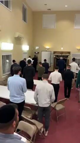 The video shows a large, rectangular room with a red carpet. About two dozen men are standing and praying towards what appears to be an altar or elevated stage at the far end of the room. The room also contains several white folding tables and brown folding chairs. The walls are painted a light yellow. There are multiple windows along one side of the room. Some of the men are wearing black skullcaps. All the men have their backs to the camera.
