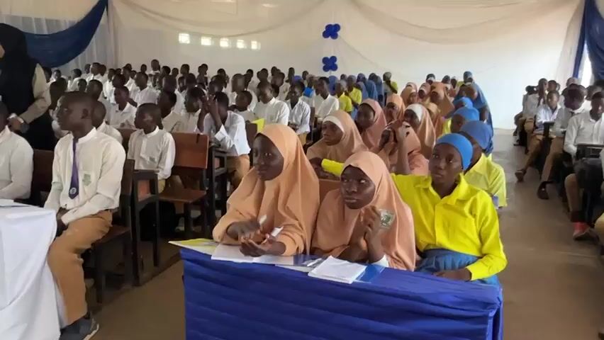 The video captures an indoor assembly or event featuring a large gathering of students, both male and female, in what appears to be a school hall in Unguwan Dosa, Nigeria. The setting is decorated with white and blue draped fabric along the walls and blue balloons.

In the foreground, several female students are seated at a table covered with a blue tablecloth. They are wearing hijabs, with some in light brown and others in blue, paired with yellow shirts. Papers and pens are visible on the table in front of them, suggesting they are engaged in note-taking or a similar activity. They appear attentive, looking towards the front of the room.

Behind the front-row students, numerous other students are seated in rows of wooden chairs. Most of the male students wear white shirts, some with ties (yellow or purple), and light brown or khaki trousers. Many female students in the background are also wearing hijabs, some in yellow shirts and others in blue attire. An adult woman in a black hijab and dark top can be seen walking through an aisle among the students.

The activity appears to be a presentation or address, with an audible speaker. The students are generally quiet and focused, observing the front of the room. The event takes place during the daytime, indicated by the lighting. No specific legible text is visible on any objects or surfaces. The scene depicts a formal educational gathering.
