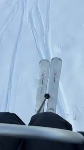 The video shows a first-person perspective of a person riding a ski lift up a snowy mountain. The person is wearing black ski pants and has skis with the brand name "Volkl" printed on them. Ski tracks are visible in the snow below, and other skiers can be seen on the slope. The ski lift consists of black chairs suspended by cables from metal towers. The sky is blue with some clouds, and the mountain peaks are covered in snow.