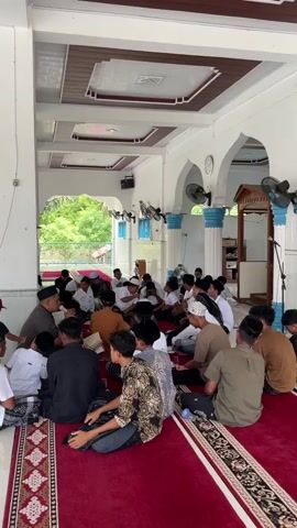 A group of young men and a few older men are gathered indoors, seated on a red carpet on the floor. They appear to be in a mosque or a similar religious building, indicated by the arched doorways, white pillars with blue trim, and a wooden structure that resembles a pulpit. The ceiling is white with decorative panels, and several fans are mounted on the walls. Some individuals are wearing white skullcaps, and others are dressed in traditional clothing. One older man, seated to the left, is gesturing and possibly leading a discussion or lesson for the seated group. Some of the young men are looking towards him, while others are interacting with each other. The overall atmosphere seems to be one of religious gathering or education.