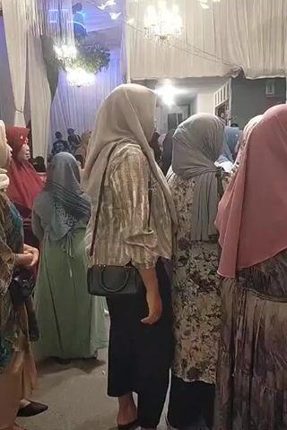 The video shows a gathering of people, mostly women wearing hijabs, in what appears to be an indoor event space. The environment is decorated with white draped fabric and chandeliers, suggesting a celebratory occasion like a wedding or party. In the foreground, a woman in a light-colored hijab and a patterned shirt is facing away from the camera, with a black crossbody bag. To her left, another woman in a light-colored hijab and a colorful patterned dress is also facing away. To the right, a woman in a pink hijab and patterned dress is partially visible. In the background, several other individuals are seated or standing, some of whom are men. The lighting is dim, with some decorative lights casting a glow. The overall atmosphere seems to be one of social interaction and event attendance.