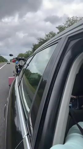 The video shows a view from inside a moving car, looking out the side window towards the back. A black SUV or similar vehicle is being filmed from the passenger side. A motorcycle is attached to a trailer hitched to the back of the vehicle. The vehicle is on a highway with other cars visible in the distance. The sky is cloudy. The video is shaky, suggesting it was filmed while the car was in motion.