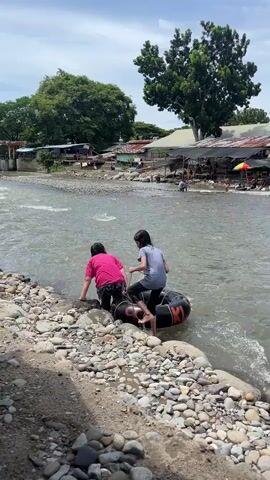 Two young girls are at the edge of a rocky riverbank preparing to go tubing. The girl on the left is wearing a grey shirt and black pants, and she is holding a rope attached to a large black inner tube. The girl on the right is wearing a pink shirt and black pants and is already seated on the inner tube, facing away from the camera. The river is flowing with moderate current, and there are small waves. In the background, there are more people, some sitting under makeshift shelters with colorful umbrellas, and trees. The sky is partly cloudy.