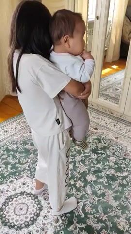 A young girl with dark hair is holding a baby in her arms. The girl is wearing a white t-shirt and white pants, and the baby is wearing a light blue shirt and light gray pants. They are standing on a rug with a green and white pattern. In the background, there is a large mirror and a window with curtains. The girl is looking towards the mirror, and the baby is looking slightly to the side.