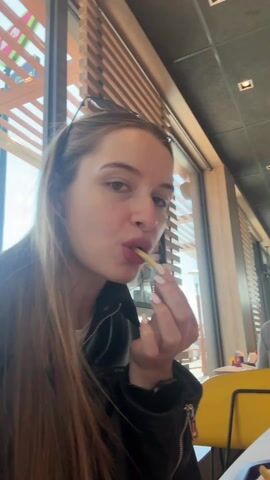 The video shows two women eating French fries at a McDonald's restaurant. The first woman has long, light brown hair and is wearing a black leather jacket. She looks directly at the camera as she eats a French fry. The second woman, who appears in the second part of the video, has long brown hair and is wearing a red and brown striped sweater. She also looks at the camera while holding a French fry and smiling. There are other people visible in the background, and the McDonald's restaurant's interior, including tables, chairs, windows, and ceiling fixtures, are visible. A McDonald's cup and French fry container are also on the table.