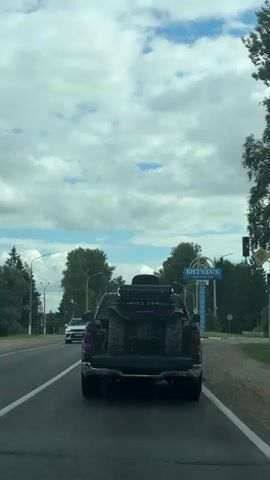 The video shows a rear view of a black pickup truck driving on a road. An ATV is loaded in the bed of the pickup truck. A white car is in front of the pickup truck, and a red taxi is to the left of the pickup truck. In the background, there is a blue sign with white text that appears to be a city limit sign, and there are trees and a traffic light visible. The sky is partly cloudy.