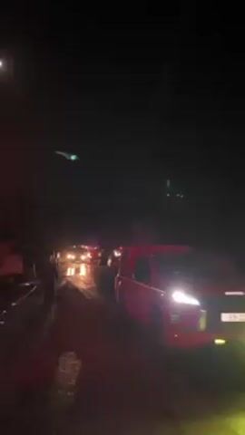 Here is a concise description of the video:

The video shows a nighttime scene where emergency vehicles are present. The road is wet, and the lights of the vehicles reflect on the surface. Several figures can be seen standing near the vehicles, possibly personnel involved in the emergency response. The sky is dark with a visible moon in the upper left, and there are some lights visible in the distance, possibly on a hilltop.