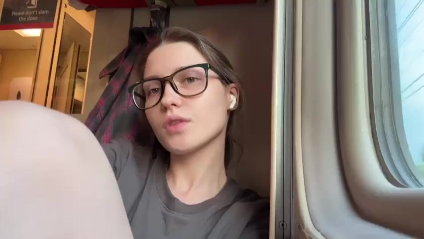 A young woman with glasses and white earbuds is sitting inside a train compartment. She is looking towards the camera with a neutral expression. A plaid jacket is hanging on the wall behind her. Sunlight is streaming in from the window, casting a warm glow on her face. The train appears to be in motion, with the scenery outside the window blurred.