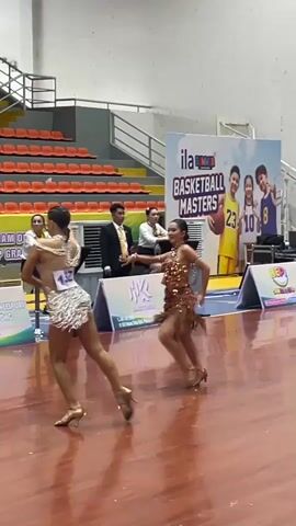 The video shows a dance performance on a wooden floor. There are two dancers in the foreground, wearing sparkly, short-skirted costumes with heels. They are moving in sync with each other. In the background, there are bleachers with orange seats, a banner that says "ILA SPORTS BASKETBALL MASTERS," and several people standing and sitting. One person is wearing a black suit, and others are in casual attire. The environment appears to be an indoor sports arena.
