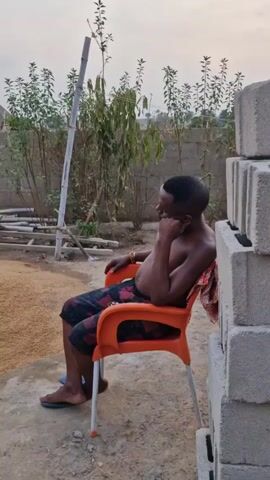 The video shows an outdoor construction site, with three men. One man is sitting on a bright orange chair, wearing patterned shorts and sandals, with his shirt off. He rests his head on his hand, appearing pensive. There are stacks of grey bricks beside him, and a mound of sand or gravel in the background. Two other men are seated on a low brick structure. One wears a light brown shirt and dark pants, and seems to be eating. The other man is shirtless, with a light blue shirt draped over his shoulders. Both men are wearing dark pants and sandals. The construction site includes cinder blocks, wooden planks, a blue and yellow portable toilet, and various building materials scattered around. A low cinder block wall surrounds the site, and sparse trees and plants border the yard. The sky is overcast.
