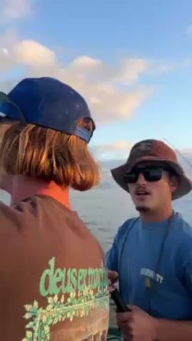The video shows a few men on a boat, likely fishing. The first man seen wears a camo bucket hat, sunglasses, and a blue Carhartt shirt. The second man wears a blue baseball cap and a shirt with green, blue, and orange graphic elements. A third man, who seems to be driving, wears a black jacket and a dark baseball cap. The camera then pans out to show the sunset over the ocean.