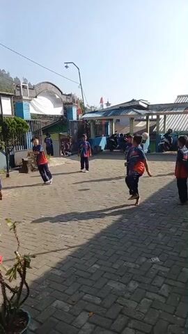 The video shows a group of young boys playing a game of volleyball outdoors in a paved courtyard. The boys are wearing matching dark blue shirts with orange and red accents and dark pants. The environment is sunny with a clear blue sky. In the background, there are buildings, a fence, parked motorcycles, and some greenery. The boys are actively engaged in the game, hitting and passing a volleyball. One boy in the foreground is facing away from the camera, walking. Another boy in the middle ground is looking towards the camera and smiling. The scene appears to be a recreational activity taking place during the daytime.
