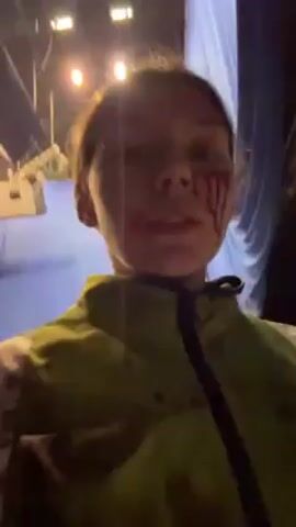 A person with fake blood makeup on their face records a selfie video as they walk down a narrow, dimly lit corridor. The person is wearing a green jacket with a black stripe. The walls of the corridor are painted light gray, and there are pipes and lights running along the ceiling. The person walks slowly through the corridor, keeping the camera focused on their face. The lighting is dim, but the fake blood is visible on the person’s face. In the beginning of the video, some stage equipment is visible.