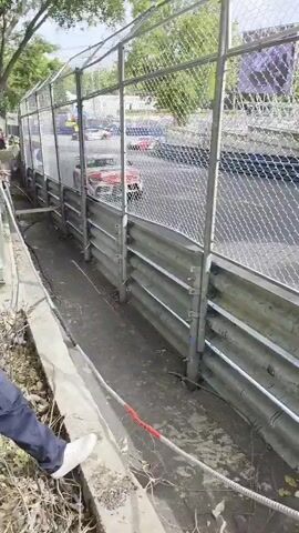 The video shows a race car, red and white with "MOTUL" branding, driving on a race track. The camera angle is from behind a metal fence, looking towards the track. Trees and foliage are visible on the right side of the track, and a white barrier runs along the track's edge. The car is the primary focus, appearing to be in motion. The overall environment suggests an outdoor race circuit with spectators or onlookers positioned behind safety barriers.