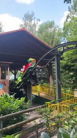A train of green and yellow roller coaster cars, designed to resemble cartoon dragons, moves along a track through a wooded area. The cars carry several passengers, some of whom appear to be children. The roller coaster is elevated, with support beams visible beneath the track. In the background, there is a wooden structure with a red roof, and a sign that reads "Ausgang," meaning "Exit" in German. Lush green foliage surrounds the roller coaster track. The sky is visible above, with some clouds.