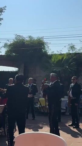 A group of six mariachi musicians in black suits with silver trim play in an outdoor courtyard. The musicians are standing on a brick patio. One plays the guitar, two play trumpets, two play violins, and one plays the vihuela. A low wall is visible in the background, covered in greenery. The sky is clear and blue. The musicians are facing a small group of seated people. Tables with white cloths and yellow chair covers are visible in the background.