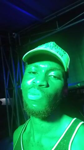 A man wearing a white and red baseball cap with the words "Original The Black" is recording a video of himself. The room is dark but the man is lit in red light, alternating to green briefly. The man is wearing a tank top with white straps. Music is playing in the background.