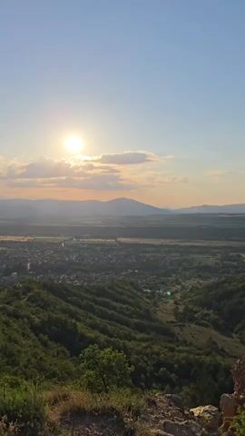 The video captures a panoramic view from a hilltop at sunset. The sun is visible in the upper left, casting warm light across the landscape. Below, a dense green forest covers rolling hills, leading to a town nestled in the valley. In the distance, mountains frame the horizon under a clear, pale blue sky. The camera slowly pans from left to right, revealing more of the vast landscape. The foreground shows some dry, rocky ground with sparse vegetation, suggesting a cliff edge. No people or animals are visible, and there are no specific actions or interactions depicted. The environment is natural and serene, with the sunset creating a peaceful ambiance.