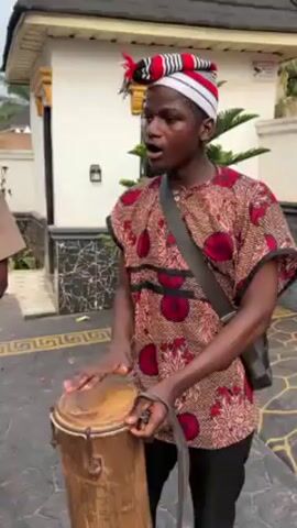 A group of young African men play music outdoors. The drummer in the foreground wears a red and brown patterned shirt, black pants, a shoulder bag, and a red and white hat. Two other musicians wear similar hats. One wears a brown "LUCKY" t-shirt and plays a drum. The other wears the same shirt as the first drummer and holds yellow musical instruments. A table with drinks and an older man sitting at it is visible in the background. The music is lively. The scene appears to be on a paved surface in front of a white house with a black roof. Security cameras are visible on the house.