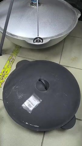 The video shows a close-up view of two large cooking pots, likely cauldrons or dutch ovens, on a tiled floor. A person's hand, wearing a striped sleeve, is seen lifting the lid off the black pot in the foreground. The black pot has a round yellow and red sticker with text on the inside. The lid is then placed down. The silver pot is visible in the background. The lighting is moderate, and the environment appears to be indoors, possibly a kitchen or storage area.