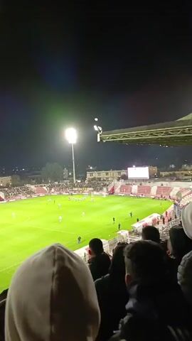 The video shows a soccer game in progress at night.  A large crowd fills the stands, with red and white banners visible. The field is illuminated by bright stadium lights. Players in red and white and blue and white jerseys compete for the ball on the green field.  Officials in black uniforms are visible along the sidelines. A digital scoreboard is visible in the distance. The camera view appears to be from the stands, slightly obstructed by the heads of other spectators.
