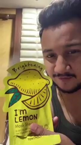 A man holds up a bright yellow TonyMoly I'm Lemon Mask Sheet. The mask packaging has a lemon drawing and the word "Brightening." He is indoors and standing in front of a door and padded wall. He has short dark hair, a trimmed beard, and is wearing a gold chain. He looks down at the mask.