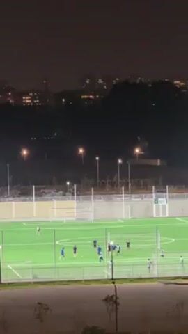 At night, a distant view shows a group of people training on a soccer field. The field is artificially lit, and is enclosed by a fence. The players are wearing blue and white uniforms. Behind the field, there are buildings and trees against the night sky. The camera zooms out slightly during the video.