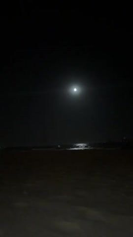 A nighttime scene on a sandy beach with the moon in the sky. The moon is bright and reflects on the water, creating a luminous path across the ocean. The beach is dark and appears empty. There is a faint glow in the distance on the right side of the frame, suggesting some man-made lights or structures. The overall atmosphere is serene and quiet.