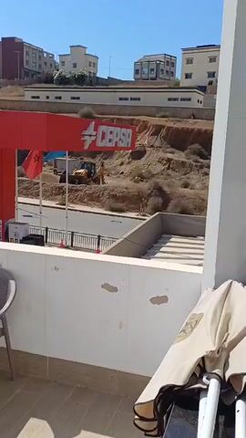 The video shows a drone shot panning across a view from a balcony overlooking a street with a CEPSA gas station. There are several buildings in the background, some under construction. On the balcony, there are chairs and a table. A yellow excavator is visible in the distance, near the gas station. The sky is clear and blue.