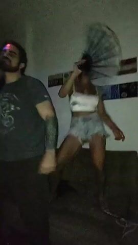 The video shows three people dancing in a room. One man is wearing a gray t-shirt with a bicycle design and black pants. A woman is wearing a white cropped top and denim shorts. Another woman is wearing a cropped light blue shirt and white shorts. They are all dancing to music, raising their hands in the air. A purple light fixture is visible on the ceiling. There is a dark couch in the background and a colorful art piece on the wall.