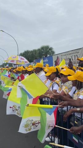 A crowd of people, mostly women, are gathered outdoors along a street, standing behind metal barriers. They are wearing uniform yellow caps and white t-shirts, many of which feature a picture of a man waving, presumably the Pope. The attendees are enthusiastically waving small flags, including the flag of Vatican City (yellow and white with a coat of arms) and the national flag of Equatorial Guinea (green, white, red, blue triangle with a coat of arms featuring a silk-cotton tree and the motto "UNIDAD PAZ JUSTICIA").

In the background, a large banner is visible with text that reads "BIENVENIDO SU SANTIDAD EL PAPA" (Welcome Your Holiness the Pope) and "A GUINEA ECUATORIAL," confirming that the event is a papal visit to Equatorial Guinea. Another part of the banner mentions "LEÓN XIV." The sky is overcast, and palm trees and street lights line the street. The atmosphere appears joyous and expectant, with many people smiling and looking towards the direction of the camera or an approaching procession. Some individuals are seen holding their phones, possibly recording the event.