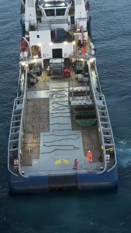 The video shows an aerial view of a supply vessel at sea. The vessel has a large open deck with ropes and some containers. Two individuals in work attire are visible on the deck. The vessel appears to be approaching a large offshore structure, possibly an oil rig, which is partially visible on the left side of the frame. The sky is overcast with a hint of dawn or dusk. The water is dark and calm. The camera angle shifts from directly above the vessel to a wider shot showing the vessel and the offshore structure.