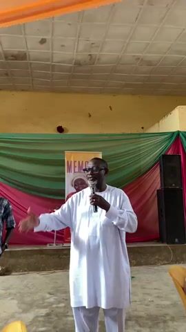 A man wearing a white outfit and glasses stands holding a microphone and speaking to a crowd. He is gesturing with his left hand. Behind him, a banner with the word "MEMORY" is visible, along with draped fabric in pink and green. Two speakers are positioned to the right of the banner. The room appears to be a hall with a checkered ceiling.
