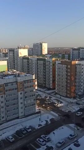 The video shows an urban landscape in daylight. It opens with a view of several multi-story apartment buildings, with snow visible on the ground and cars parked in the parking areas. The buildings have varied color schemes, including grey, orange, yellow, and green accents. The camera then pans up, focusing on a single grey building with numerous windows. Finally, the camera pans further to reveal the bright sun in a clear blue sky, with a cityscape silhouetted on the horizon. A single electrical wire is visible in the sky.
