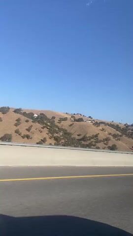 This is a POV video taken from a vehicle traveling on a highway. A concrete barrier separates the highway from a brown, hilly landscape with scattered trees and some houses. The sky is clear and blue. As the vehicle moves, a blue Amazon semi-truck drives by on the highway. Later, a black semi-truck with "CHEEMA" written on the trailer drives past. The video ends with a black car visible further down the highway.