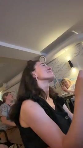 A young woman in a black top with ruffled sleeves is holding up a menu and looking at it. She has dark hair and is wearing earrings. She is seated in an indoor environment that appears to be a restaurant or cafe. Another person is visible in the background, out of focus. The lighting is dim.