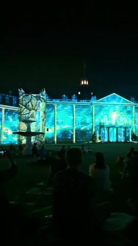 The video shows a nighttime outdoor projection mapping show on a building. The building appears to be a large, classical-style structure with columns and a central dome. The projection is a dynamic, colorful display featuring abstract patterns, possibly representing water or nature elements, in shades of blue and green. In the foreground, a crowd of people are seated on the grass, facing the building, watching the show. The lighting is dim, typical of an evening event, with the building's illuminated facade being the primary light source.