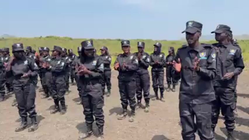 Here's a concise description of the video:

A group of individuals, likely police or security personnel, are lined up in formation outdoors. They are wearing matching black uniforms with hats and patches on their shoulders. The group is participating in some type of coordinated activity, moving their arms and hands in unison, almost like a dance or exercise routine. The background features a grassy field and a clear sky, suggesting a rural or training environment.